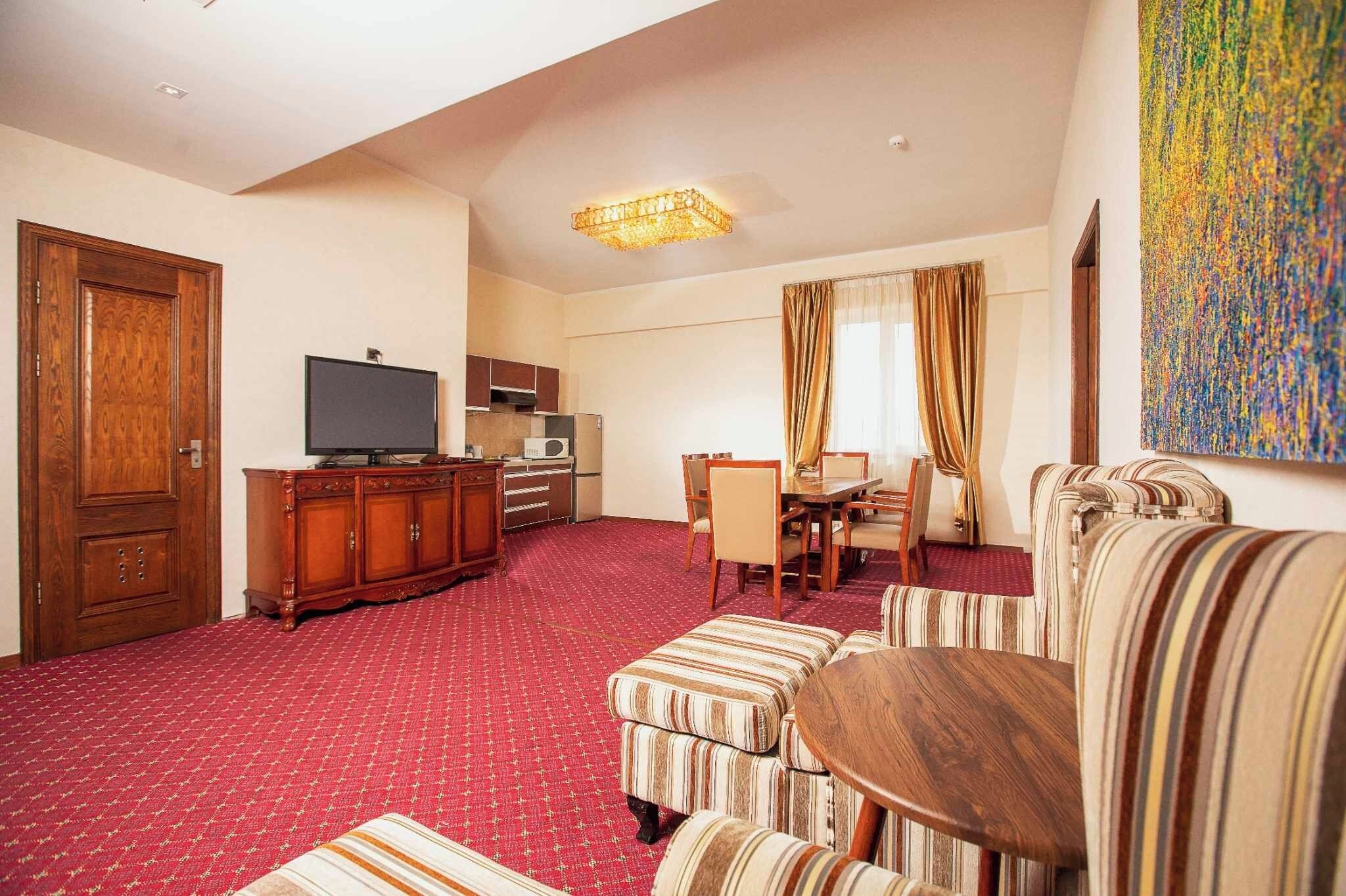 Habitación de hotel con alfombra roja, muebles de madera, televisión, kitchenette y un cuadro abstracto grande.