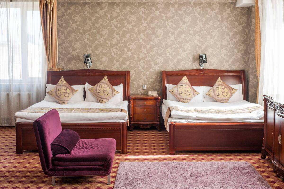 Habitación de hotel con dos camas de madera, un sillón morado, una mesita de noche pequeña y una alfombra estampada.
