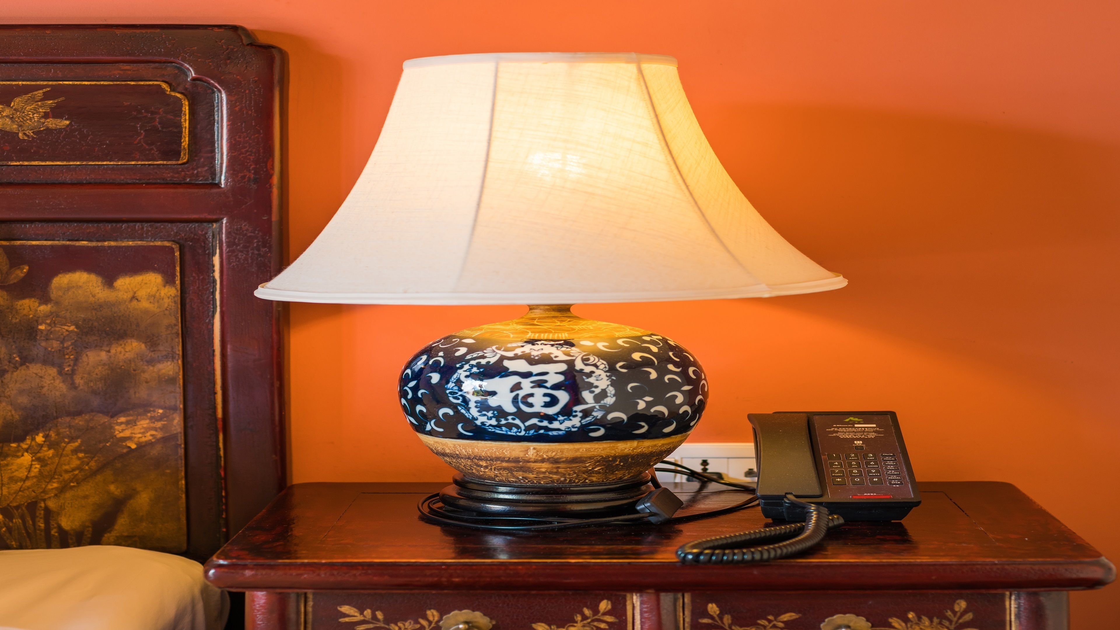 A table with a decorative lamp, a telephone, and a wooden nightstand against an orange wall.