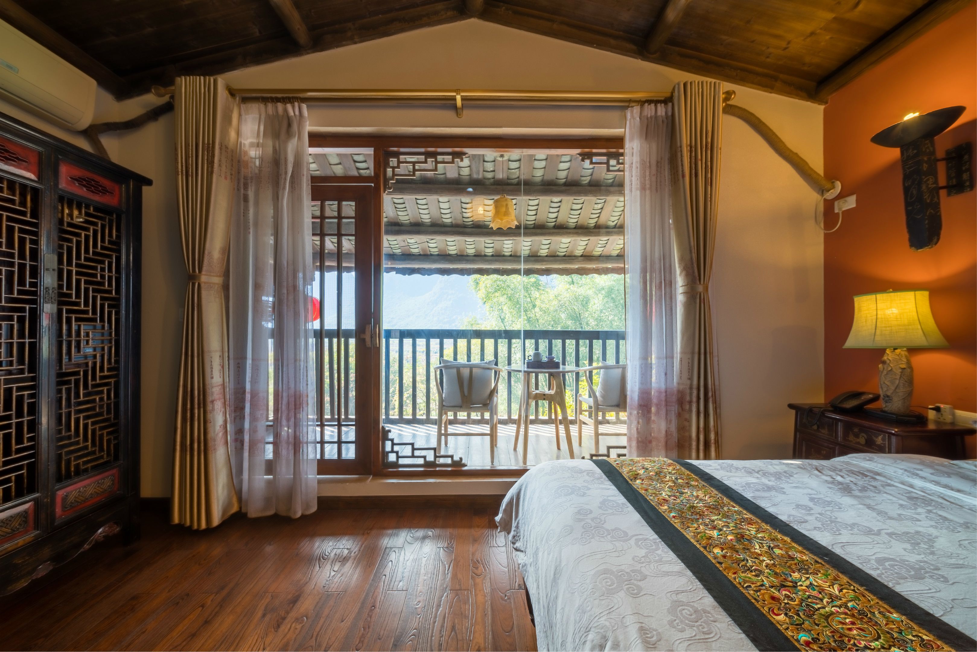 A traditional room with a wooden floor, a bed with a patterned bedspread, a balcony with a table and chairs, and a decorative wall hanging.