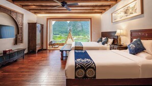 Ein Zimmer mit zwei Betten, Holzboden, Deckenventilator, großem Fenster mit Blick auf Grünflächen und einem Holzschrank.