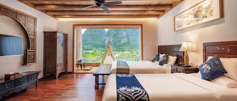 Una habitación con dos camas, suelo de madera, ventilador de techo, un ventanal con vistas a la vegetación y un armario de madera.