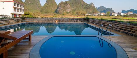 A swimming pool with a wooden deck, surrounded by mountains and a building in the background.