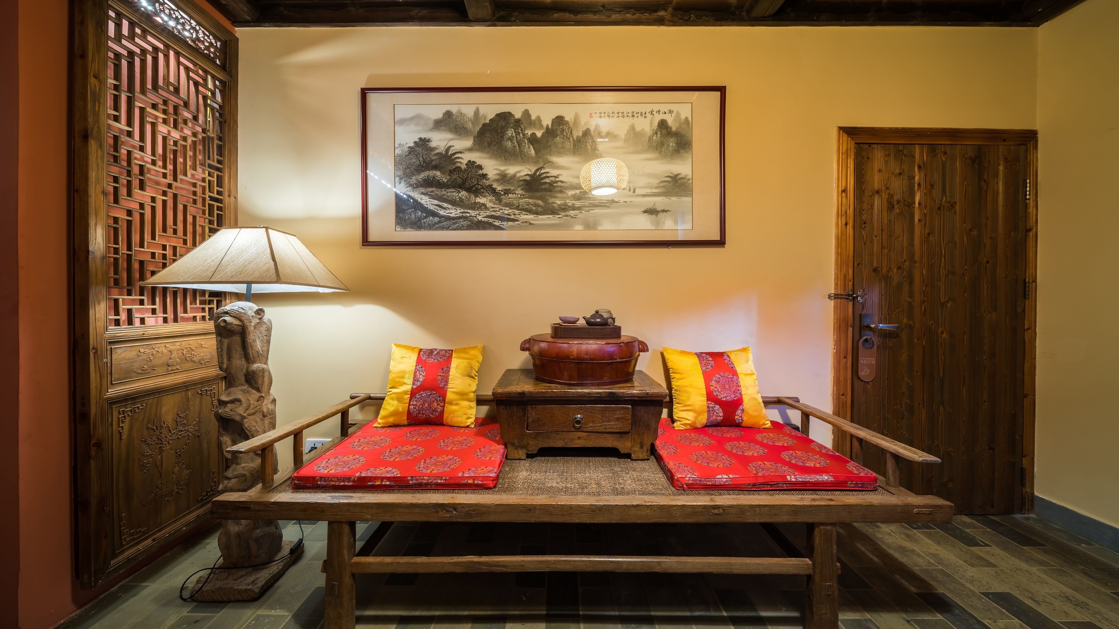Una habitación tradicional con una mesa de madera, dos almohadas rojas, un armario de madera, un cuadro enmarcado y una puerta.
