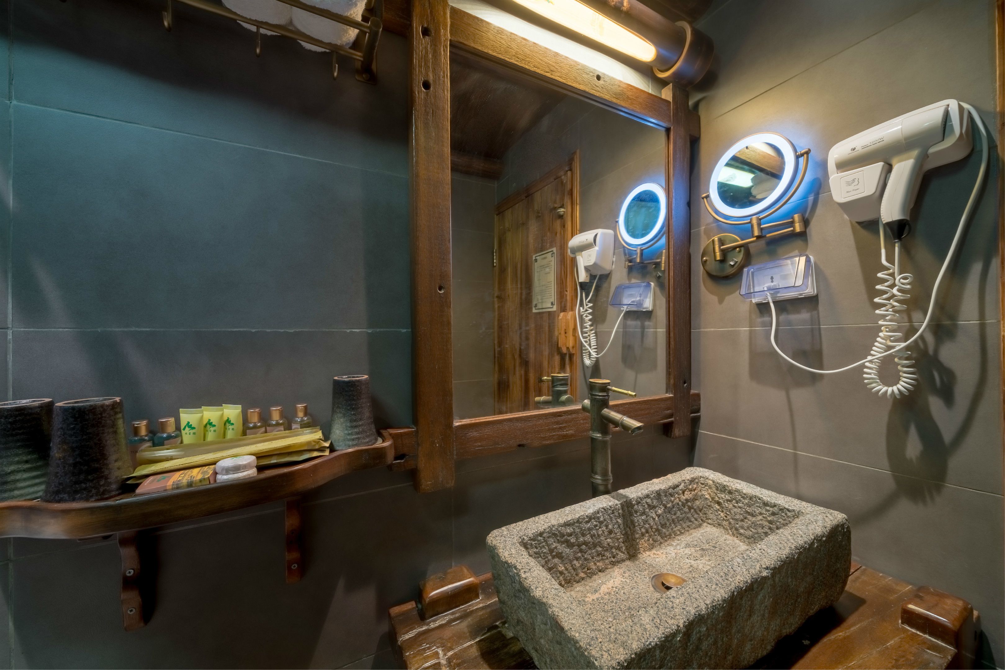 A bathroom with a stone sink, a wooden shelf with toiletries, a mirror with a round light, and a hairdryer mounted on the wall.