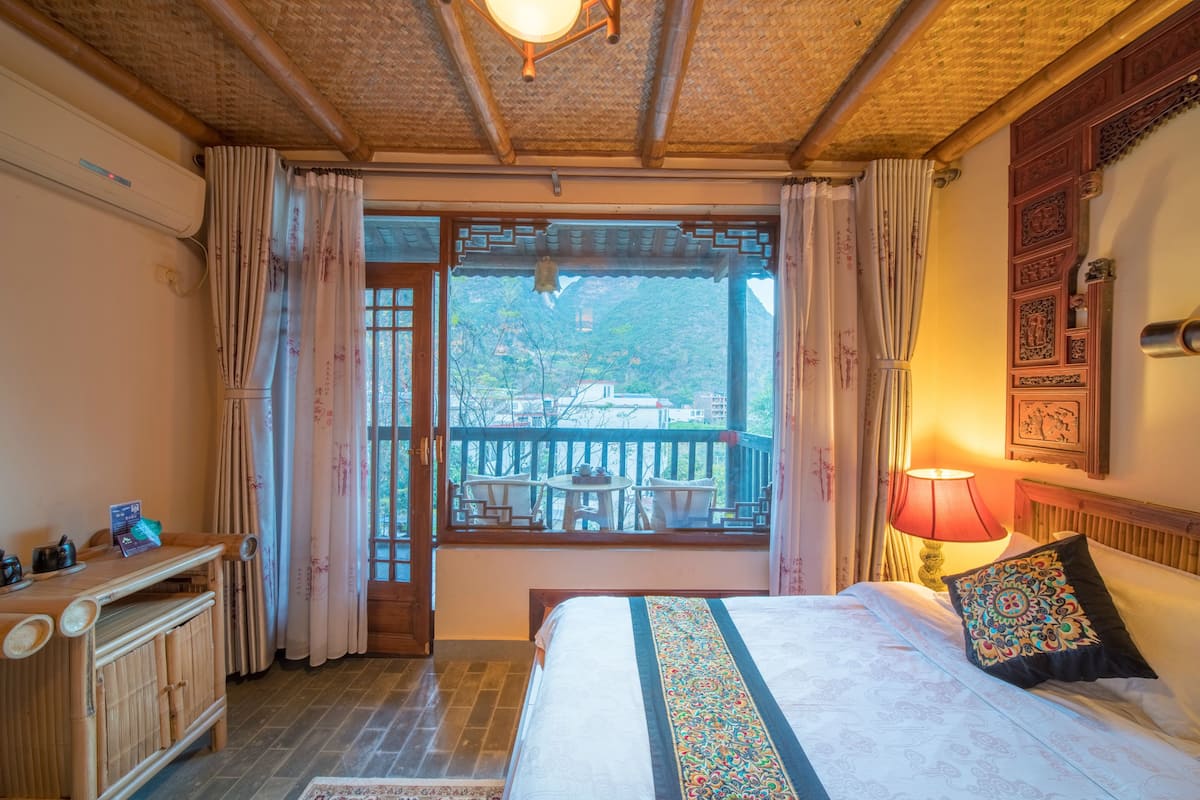 A traditional room with a bed, a wooden desk, a balcony with chairs, and a view of the outside.