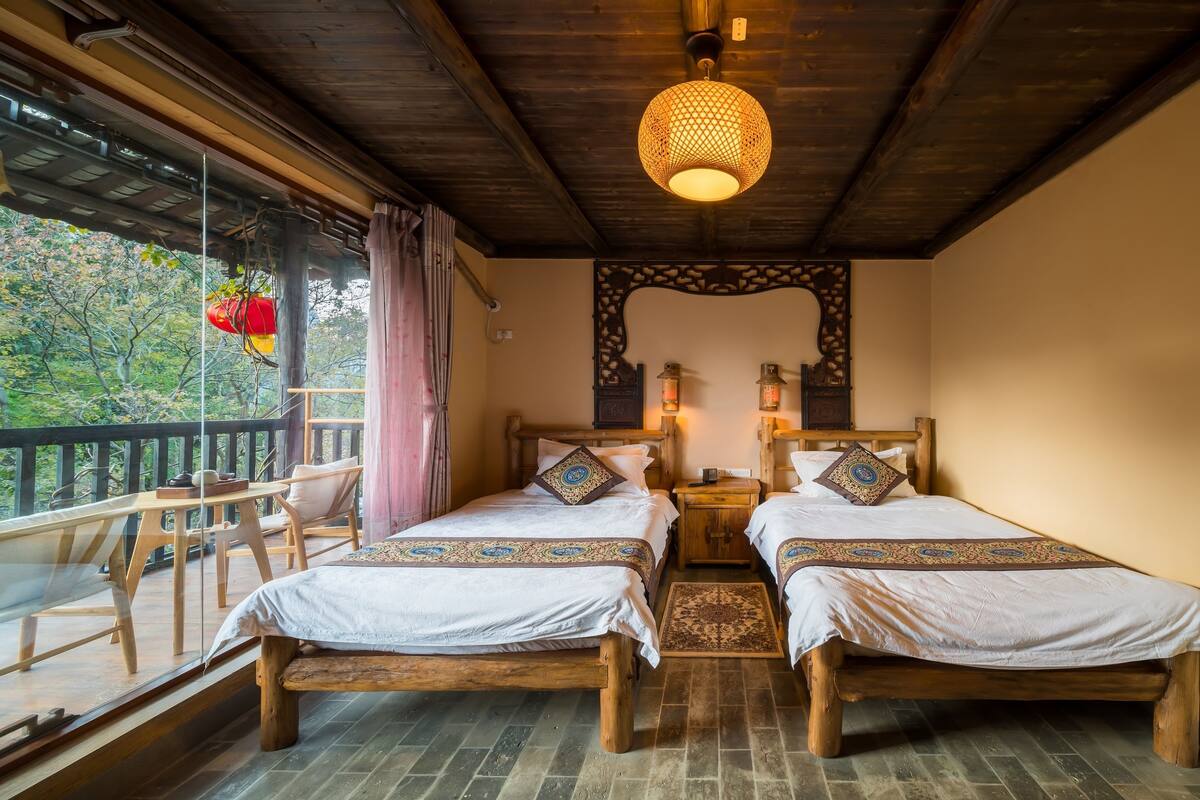 A room with two wooden beds, a wooden ceiling, a hanging light fixture, a balcony with a chair, and a view of trees.