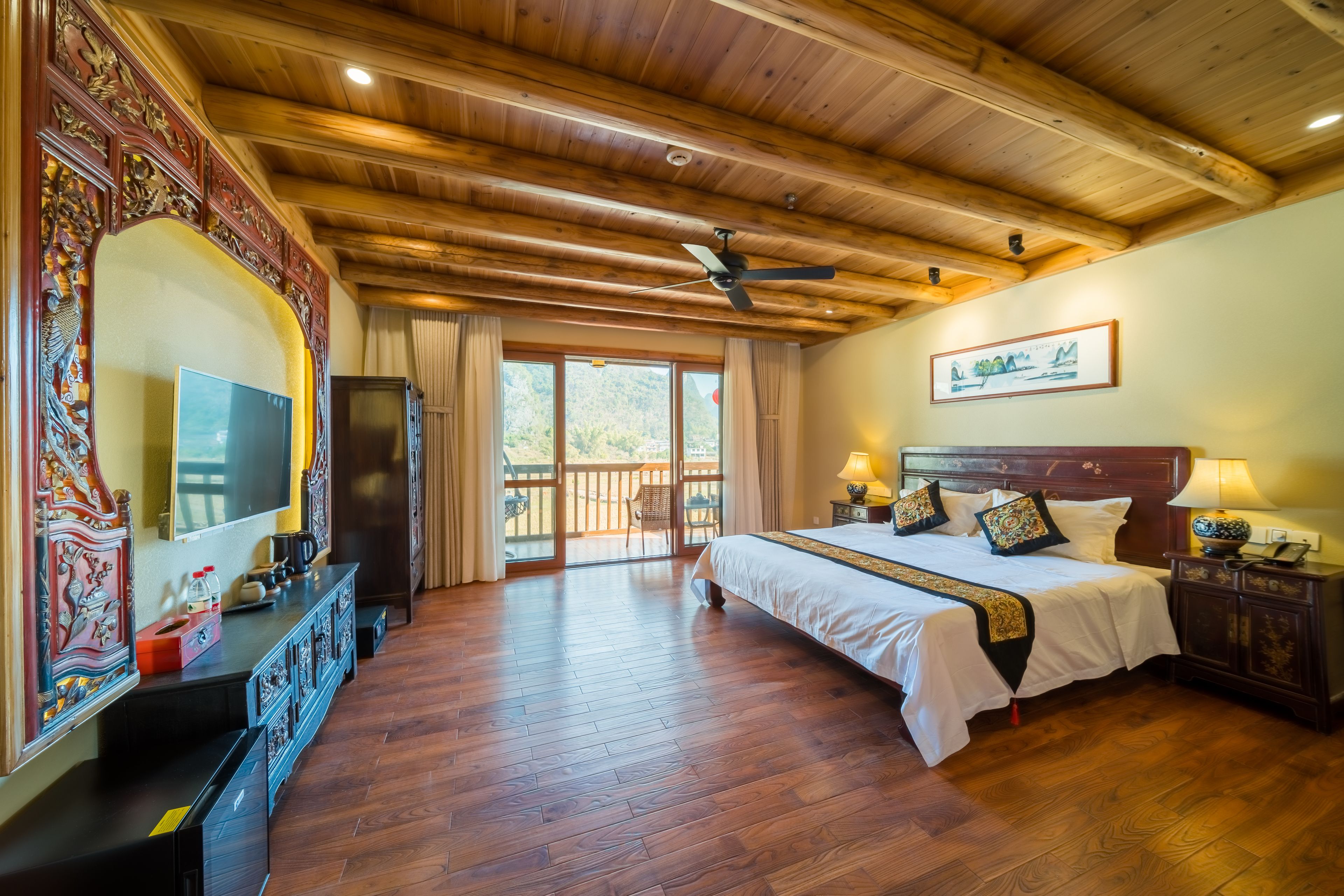 A spacious bedroom with a large bed, wooden ceiling, a television, and a balcony with a view of the outdoors.