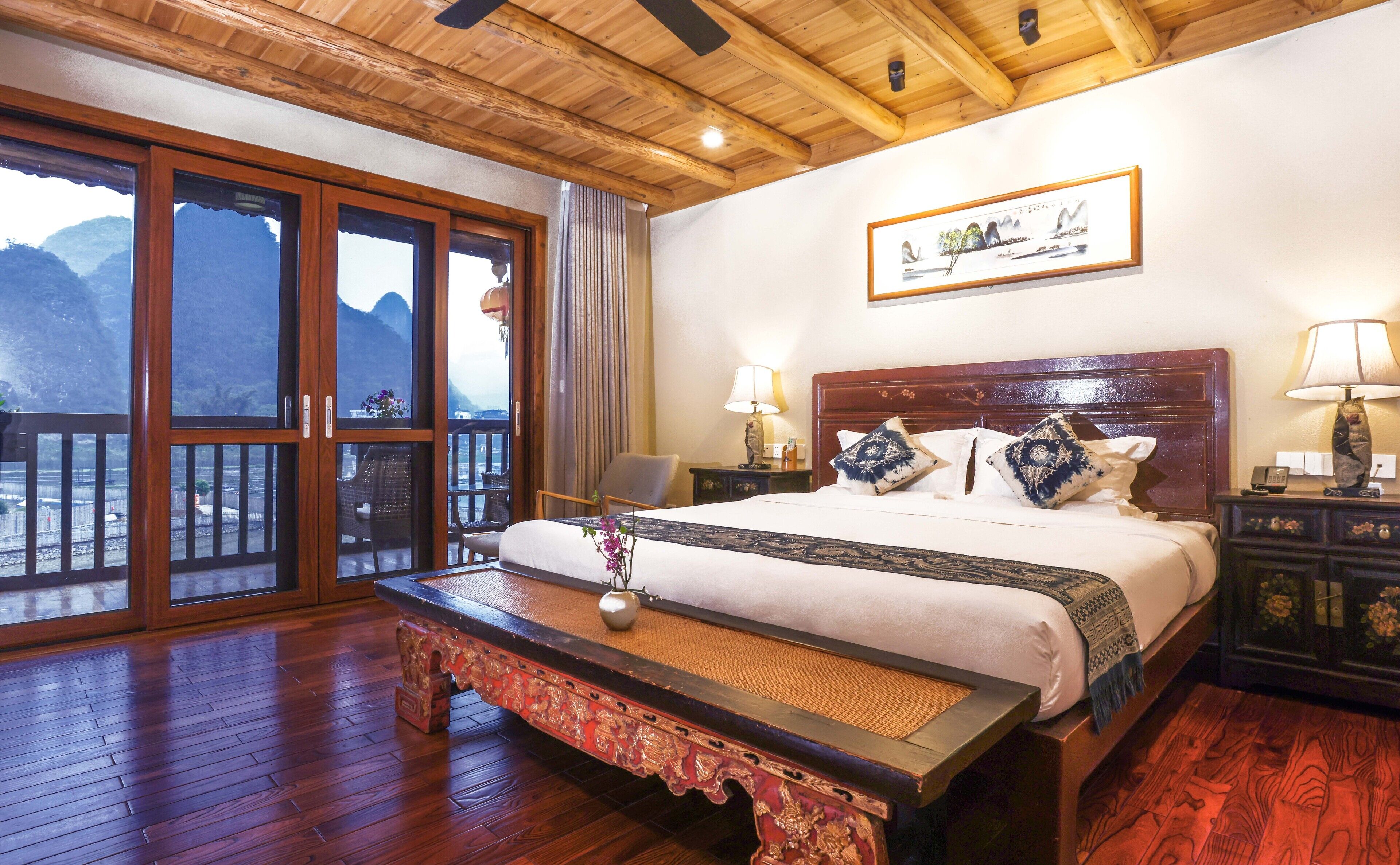 A hotel room with a large bed, wooden floor, a bench, a view of mountains through the windows, and a painting on the wall.