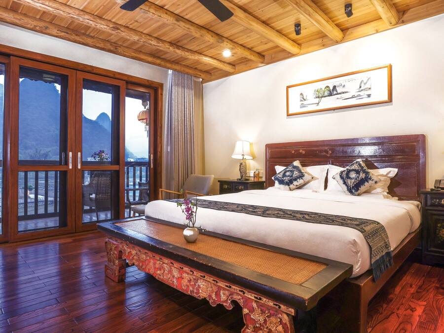 A hotel room with a large bed, wooden floor, a bench, a view of mountains through the windows, and a painting on the wall.