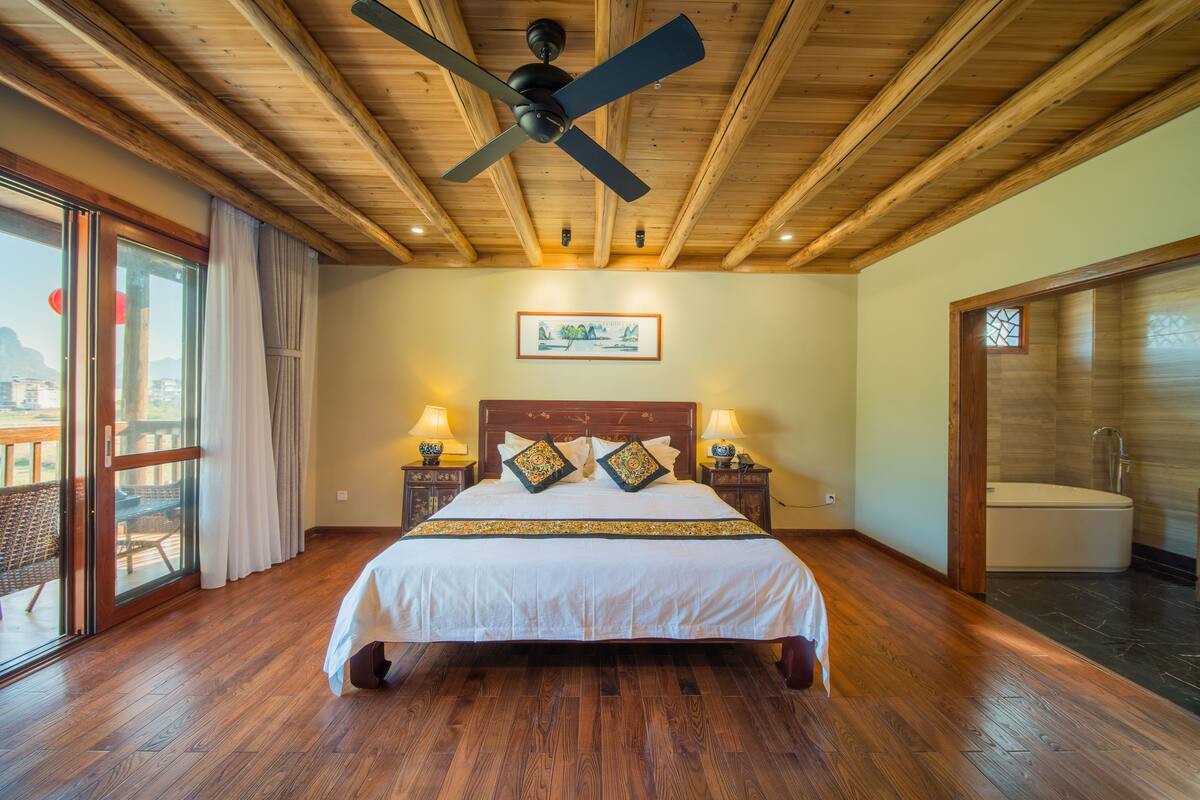 A bedroom with a wooden ceiling, a bed with white bedding, two nightstands with lamps, a painting on the wall, and a sliding glass door to a balcony.