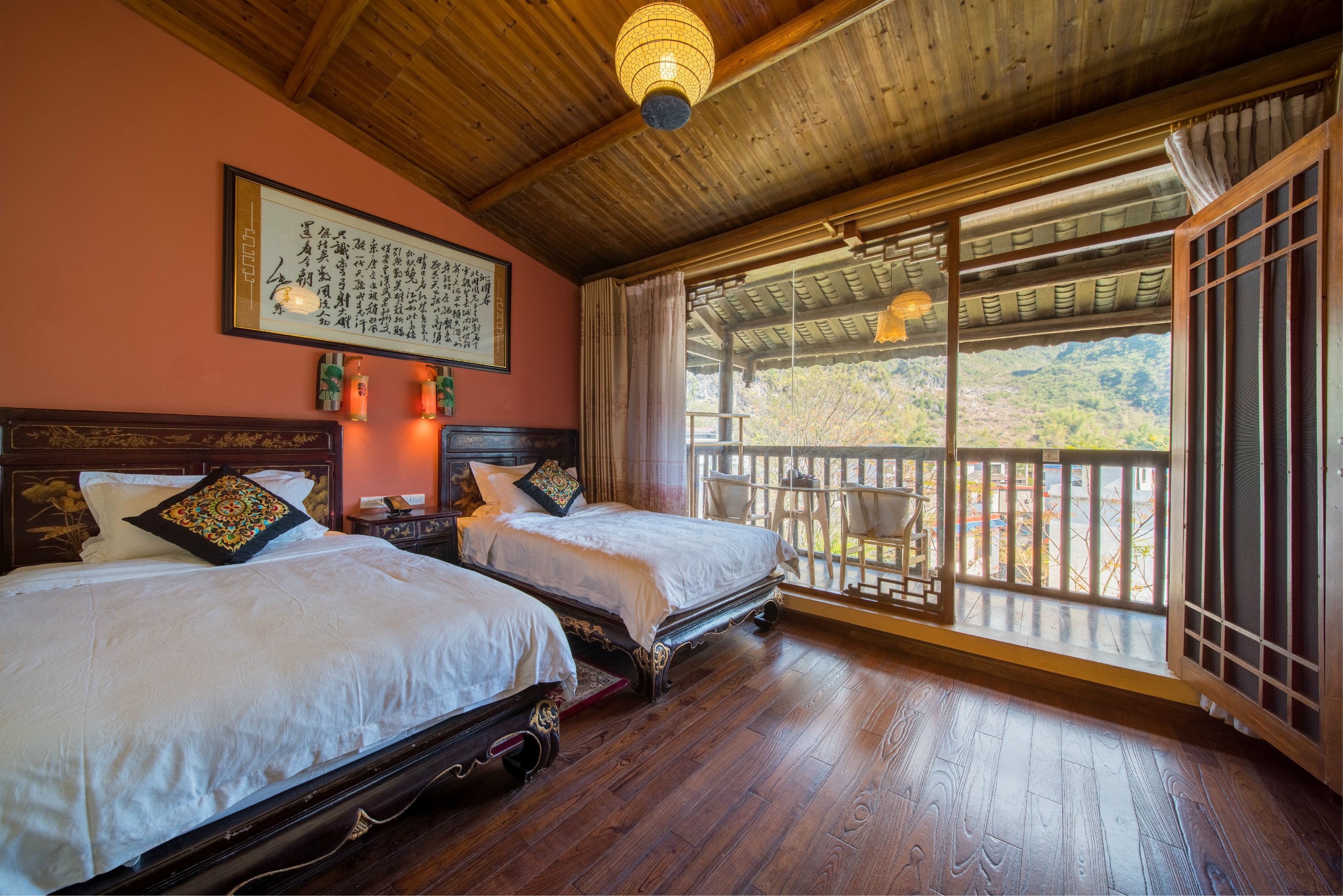 A traditional room with two beds, wooden flooring, a ceiling fan, and a view of a mountainous landscape through a large window.