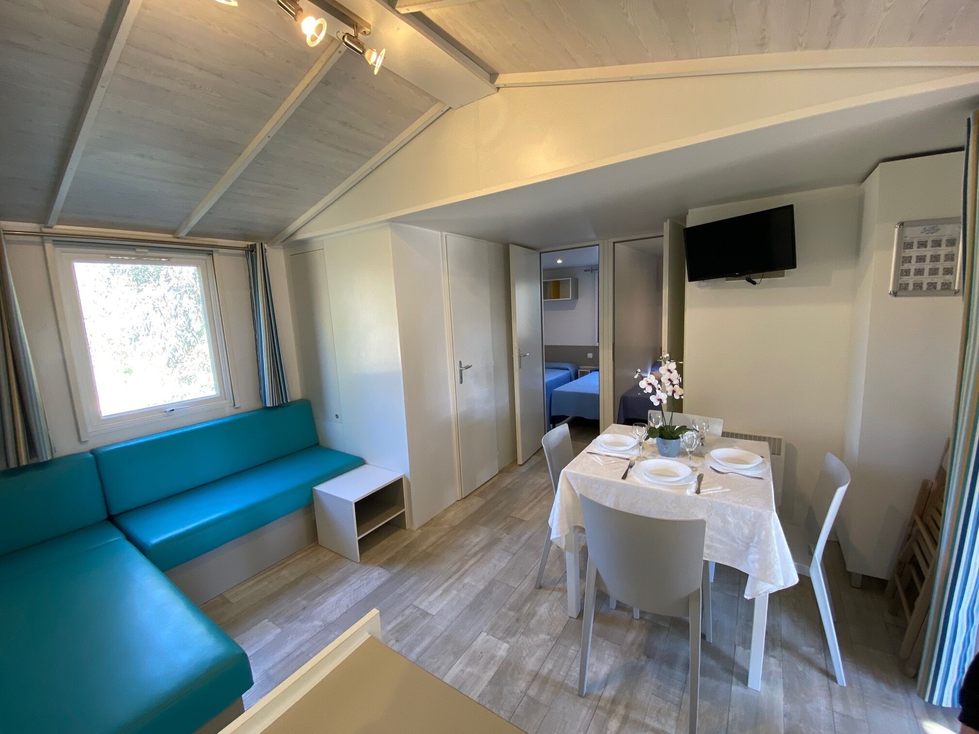 Una habitación compacta con un sofá color turquesa, una mesa de comedor lista para cuatro personas y un televisor montado en la pared.