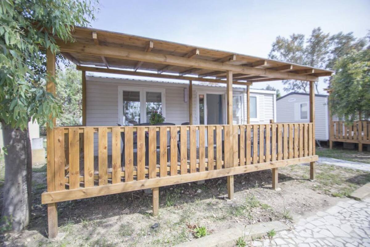 Eine Holzterrasse mit einer Pergola, ein weißes Gebäude im Hintergrund und ein Baum zur Linken.