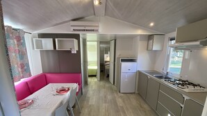 Een compacte kitchenette met een koelkast, fornuis en gootsteen. In de aangrenzende woonkamer staat een roze bank en een eettafel.