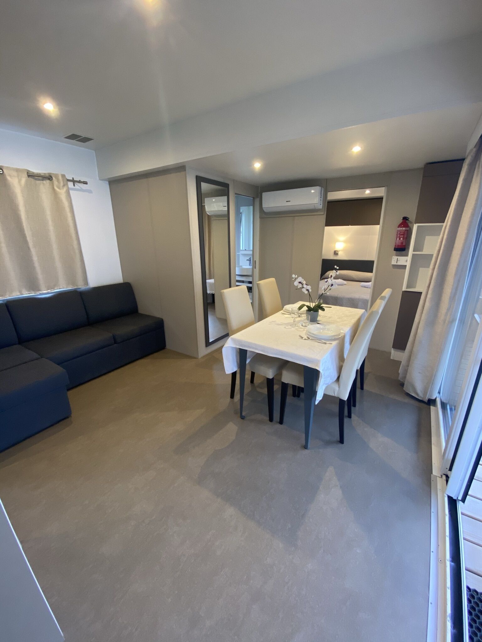 Una sala de estar moderna con un sofá azul oscuro, una mesa de comedor con sillas blancas y una cocina visible a través de un vano abierto.