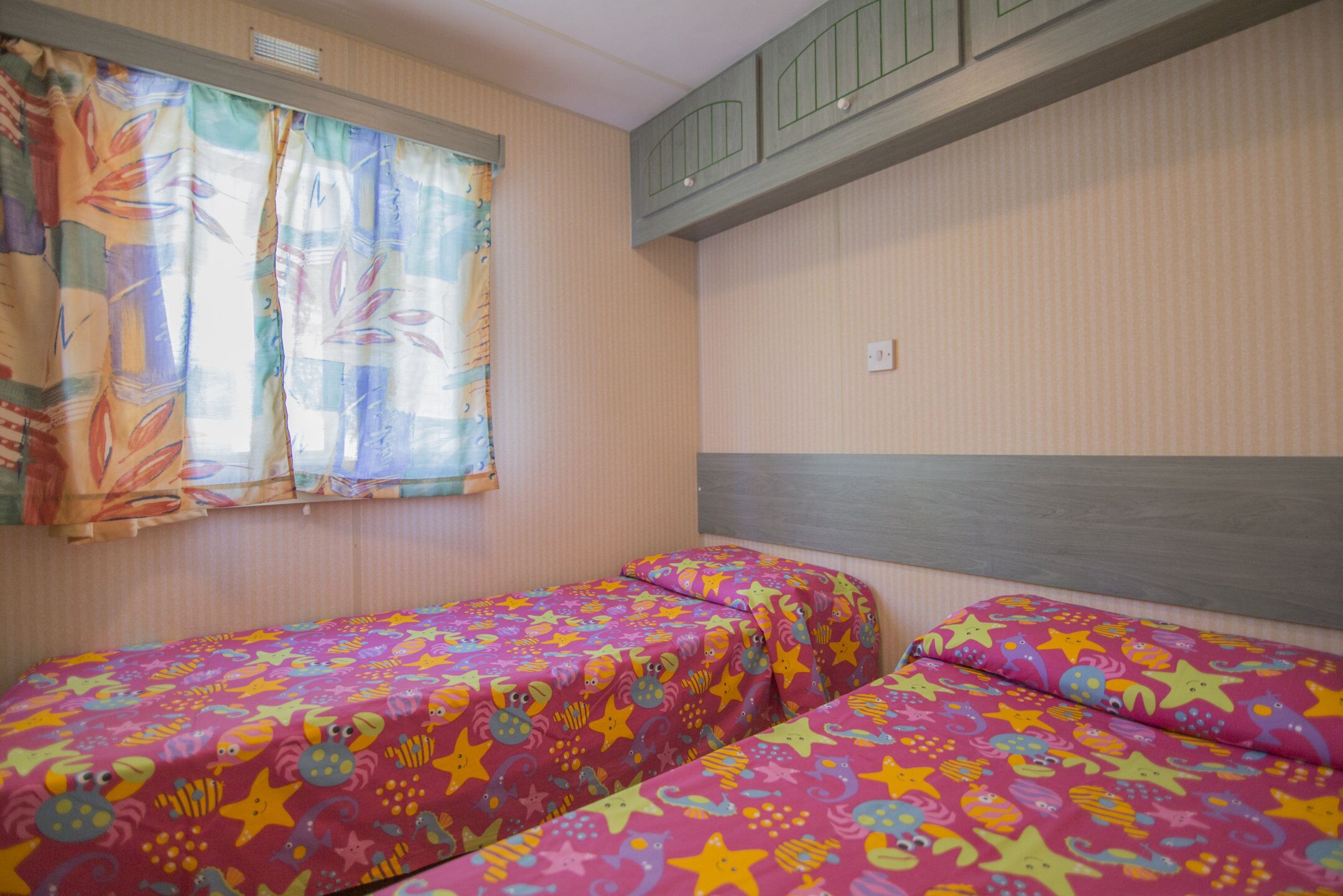 Ein Zimmer mit zwei Betten, die mit farbigem Sterndeckenmuster bezogen sind, einem Fenster mit Vorhängen und einem Lichtschalter an der Wand.