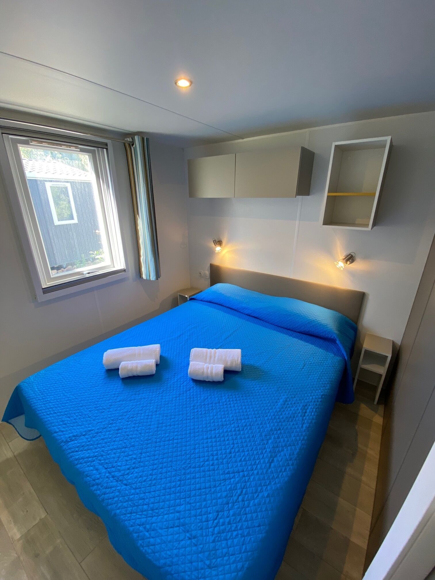 A bedroom with a blue bedspread, two rolled towels, a window with curtains, and a wall-mounted shelf.