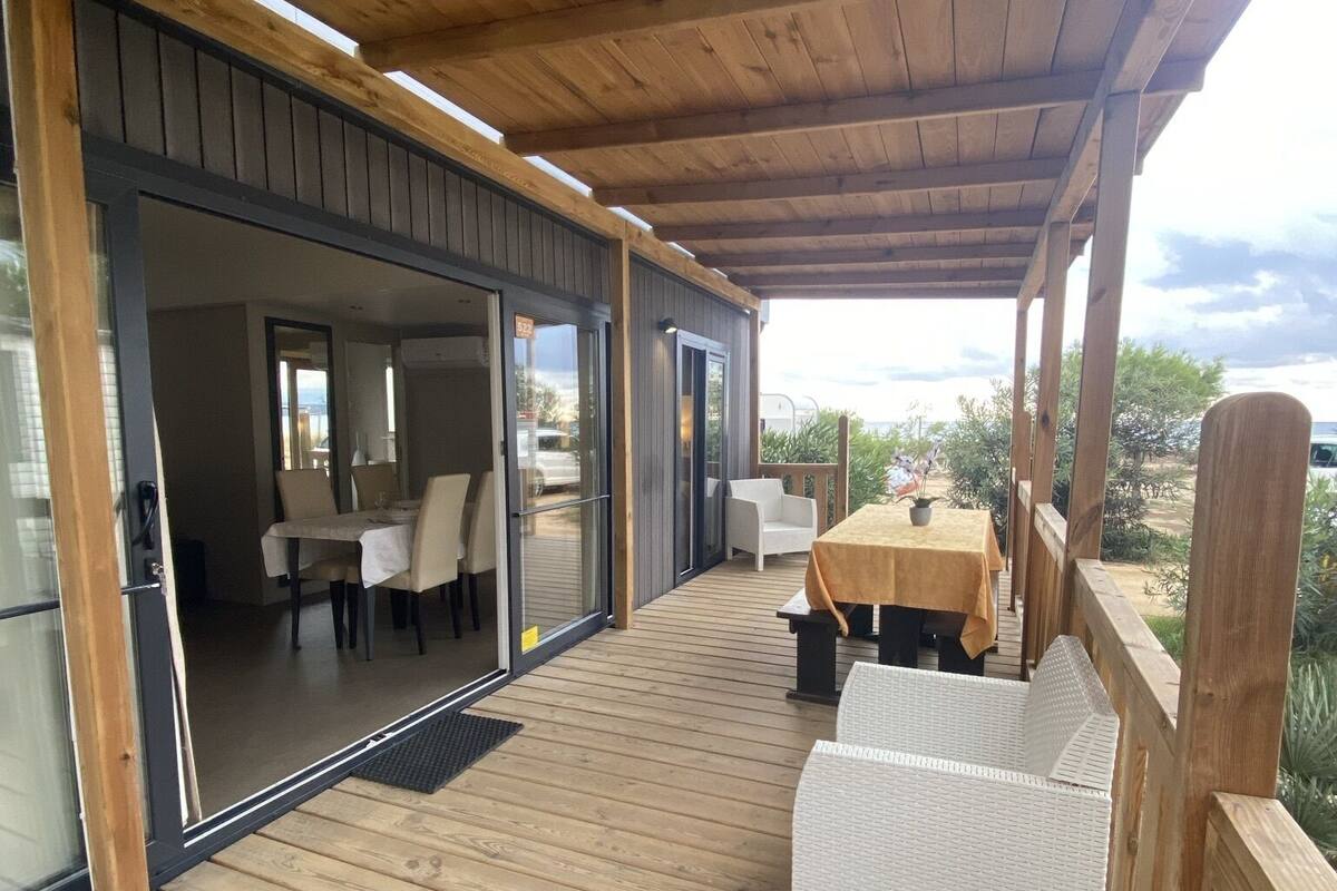 Eine überdachte Terrasse mit Holzboden, Sitzgelegenheiten im Freien und bodentiefen Glasfenstern, die zu einem Innenbereich mit Essbereich führen.