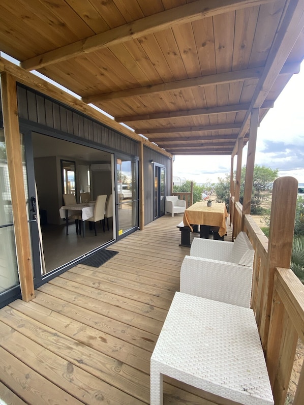 A covered deck with wooden flooring, outdoor seating, and sliding glass doors leading to an interior dining area.