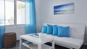 A modern living room with a white sofa, blue cushions, a white coffee table with books and a decorative item, a wicker ottoman, and a large window with blue curtains. - Beach Cabanas (Ermionida)