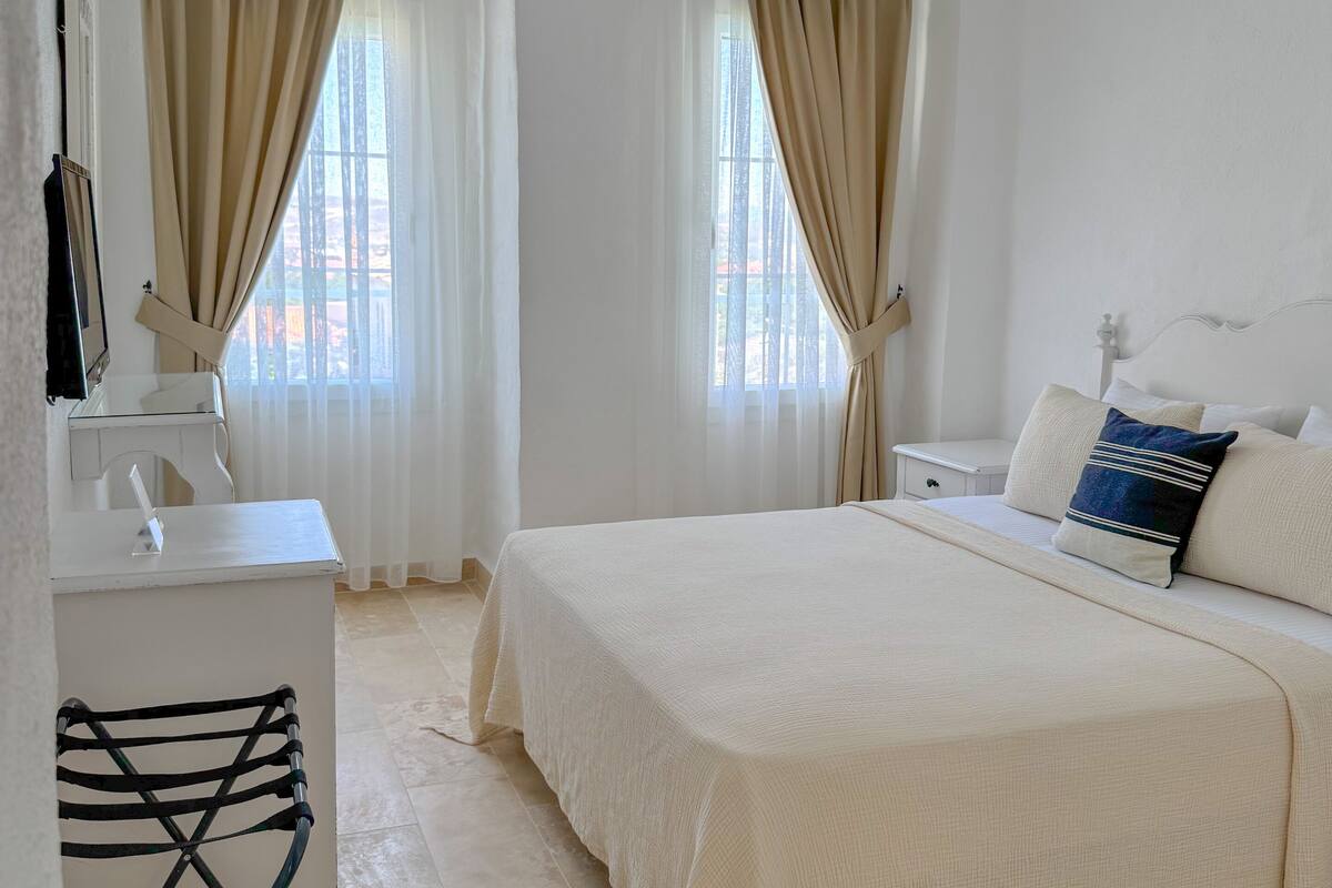 A clean, white room with a bed, a desk, and two windows with beige curtains.