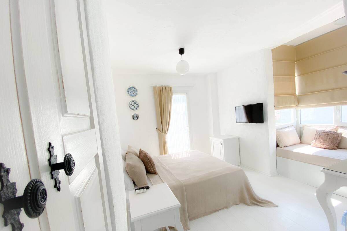 A bright, white room with a bed, bedside table, and a TV mounted on the wall.