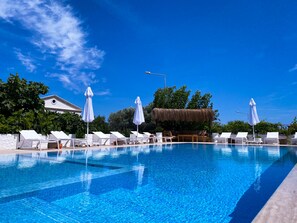 En klarblå pool med vita solstolar och parasoller, omgiven av grönska och en byggnad med tak av halm.