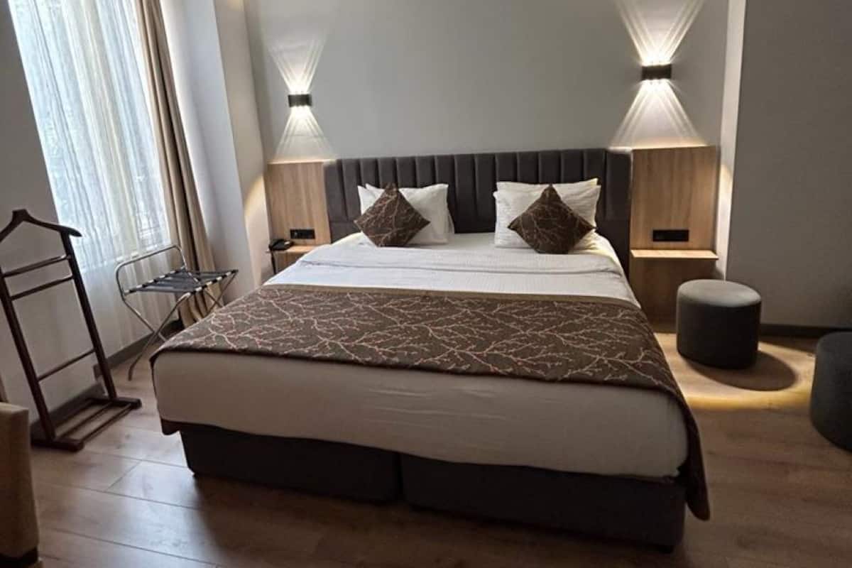 A modern hotel room with a large bed, two bedside tables with lamps, a wooden floor, and a window with curtains.