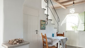 A dining area with a table set for two, a wooden chair, a decorative wall hanging, and a rustic shelf with a pile of wood. - Skaris Homes (Tinos)