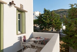 Un balcón con mesa y silla blancos, una ventana con vistas a una colina y árboles, y un muro de piedra al fondo.