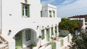 A white building with green doors and windows, a small balcony, and potted plants. - Skaris Homes (Tinos)