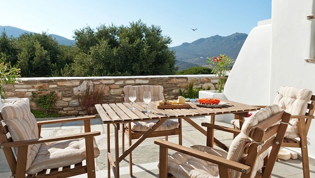 A wooden outdoor dining area with a stone wall, white walls, and a view of mountains. - Skaris Homes (Tinos)