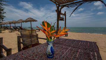 A beach setting with a table covered by a patterned tablecloth, a vase with orange flowers, and a beach umbrella in the background.