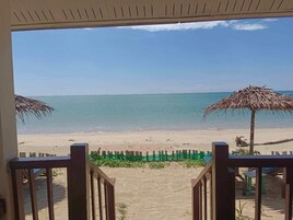 A beach view from a covered area with a wooden railing, a thatched umbrella, and a sandy path leading to the shore.