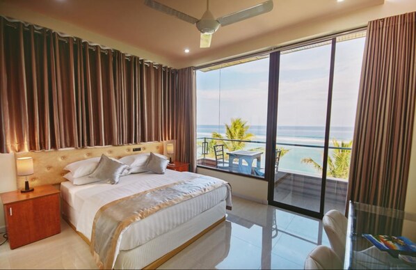 A hotel room with a large bed, a nightstand, a desk with a chair, and a balcony with a view of the ocean.