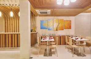 A restaurant with wooden ceiling beams, a central dining area with tables and chairs, and wall art depicting trees. - Lonuveli (Hulhumalé)