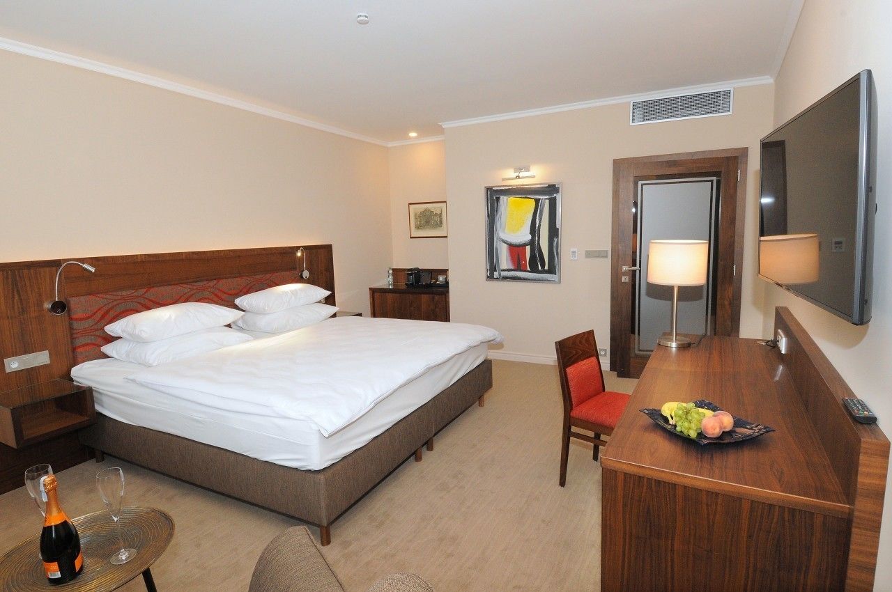 A hotel room with a large bed, a desk with a chair, a television, and a small table with a bottle and glasses.