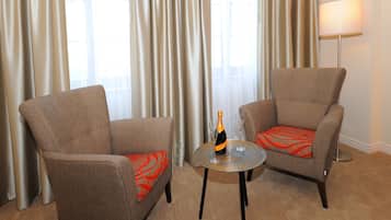 Dos sillones con cojines rojos, una botella de champán y dos copas sobre una mesita en una habitación con cortinas y una lámpara.