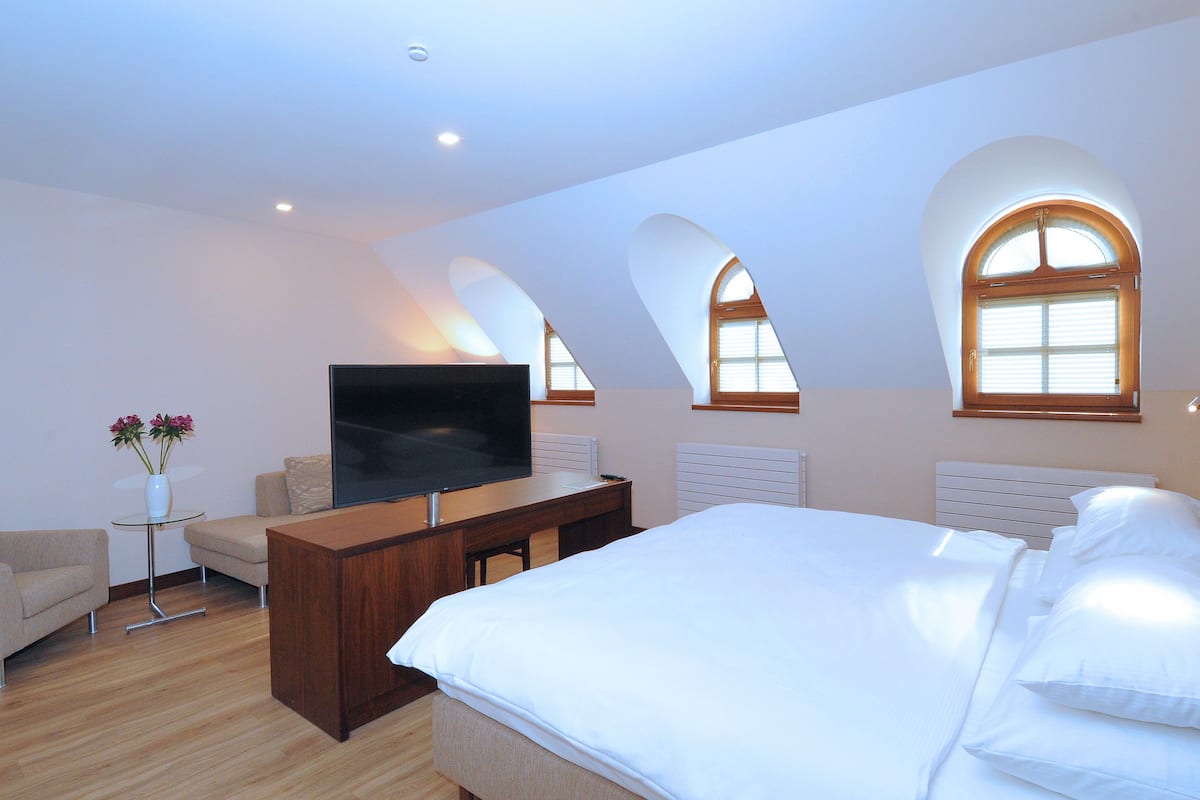 Un dormitorio con una cama grande, un televisor sobre un soporte de madera, una mesita con flores y ventanas arqueadas.