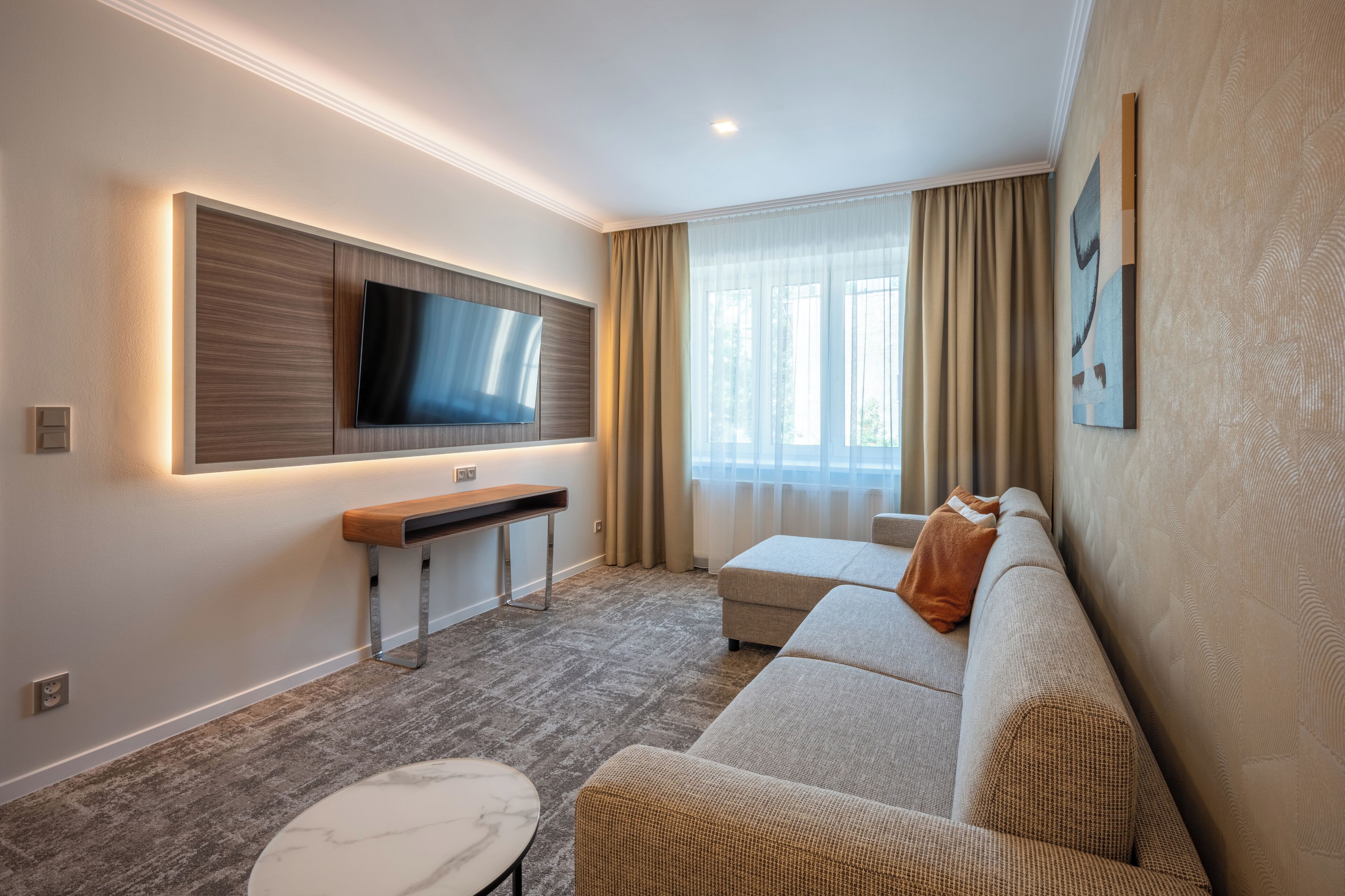 A modern hotel room with a flat-screen TV mounted on the wall, a sofa, a small table, and a window with curtains.
