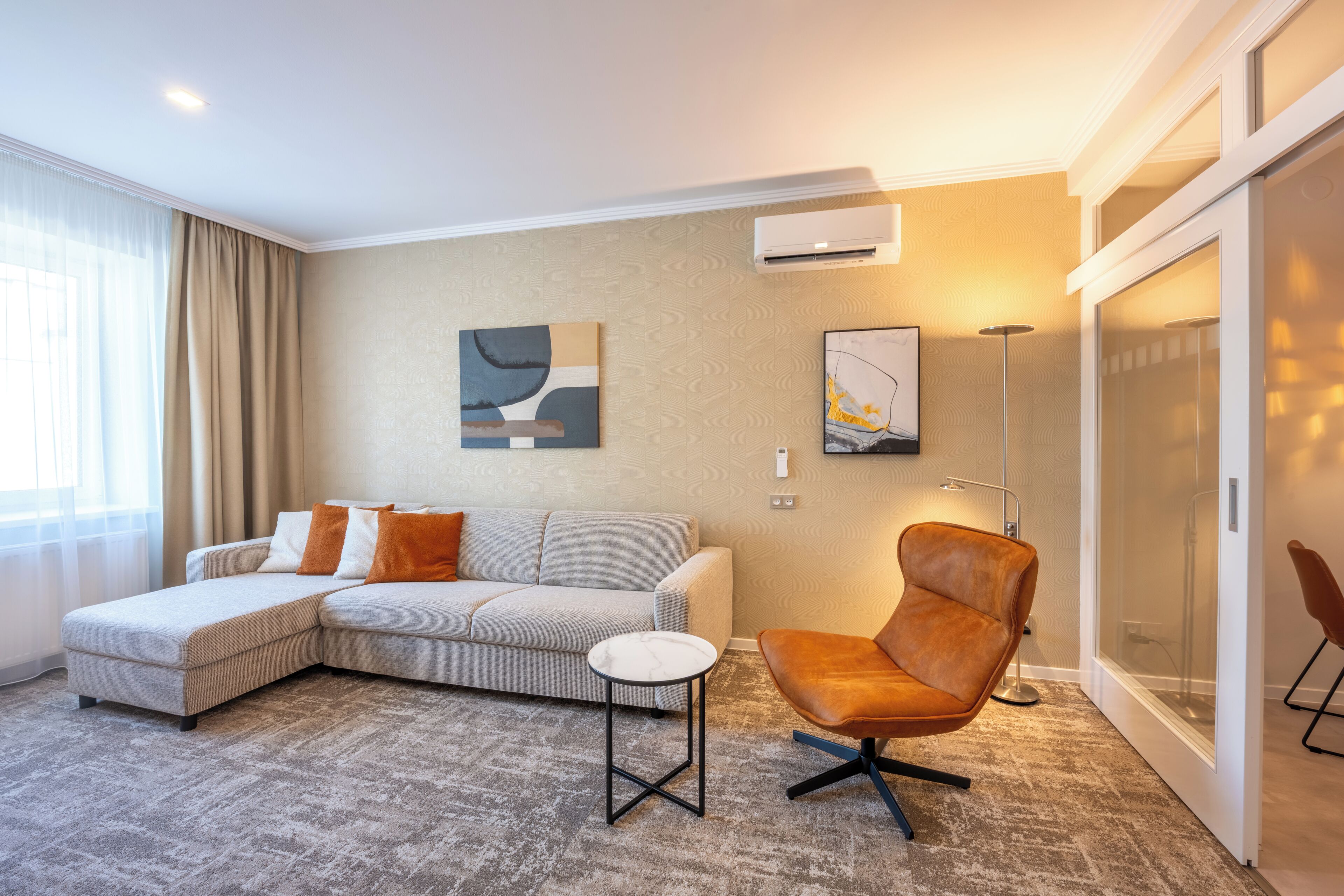 Un salon moderne comprenant un canapé d’angle, un fauteuil, une petite table d’appoint et une grande fenêtre avec des rideaux.