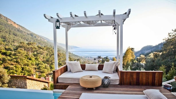 Eine Holzterrasse mit weißem Pergola, eine Lounge-Ecke mit Kissen, ein runder Tisch aus Bogenwerk und Blick auf eine gebirgige Landschaft.