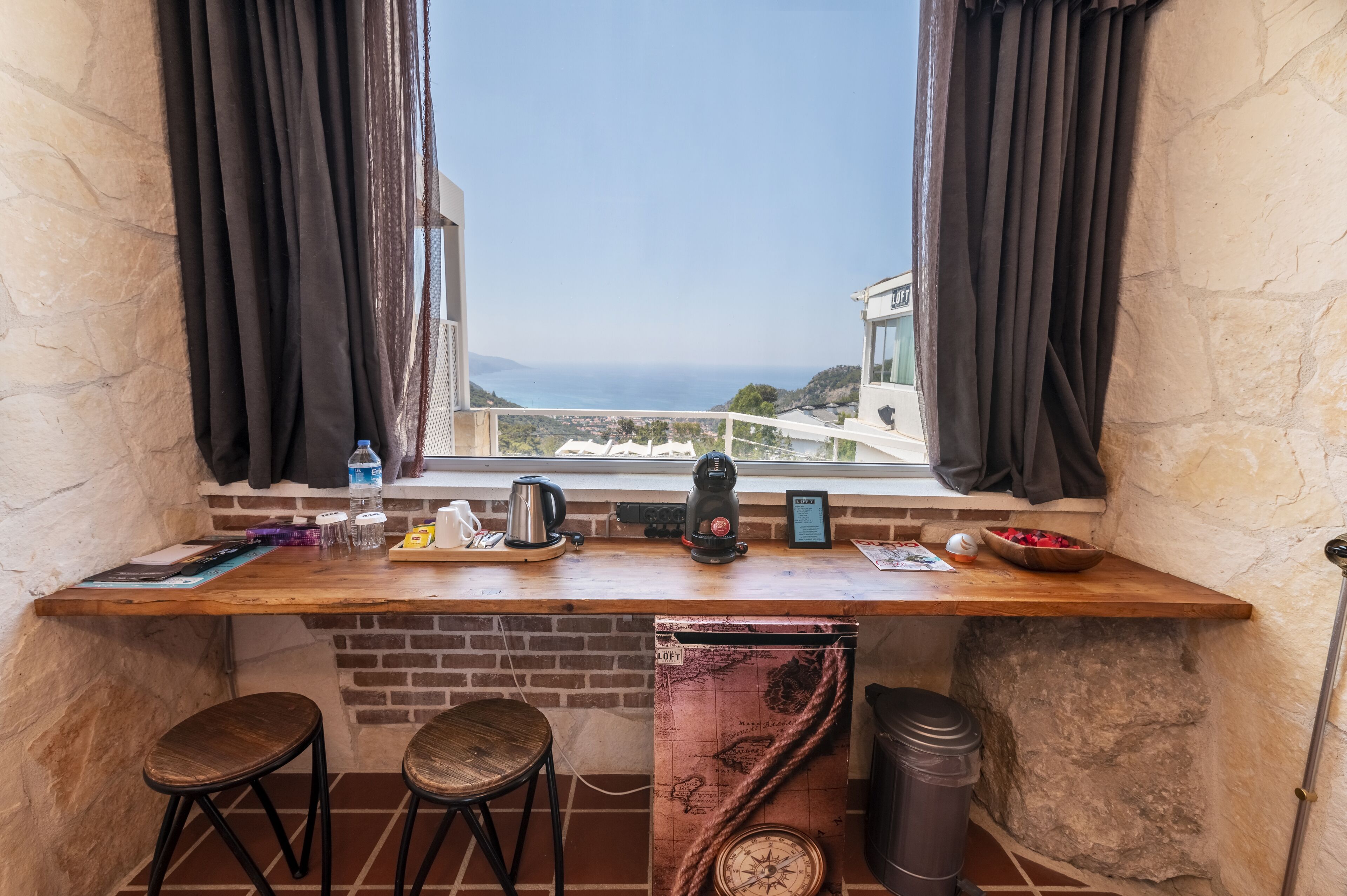 Un pequeño rincón de cocina con encimera de madera, dos taburetes, una ventana con cortinas y vistas al mar.