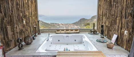 A bathroom with a large bathtub, wooden benches, and a view of a coastal town through a large window.