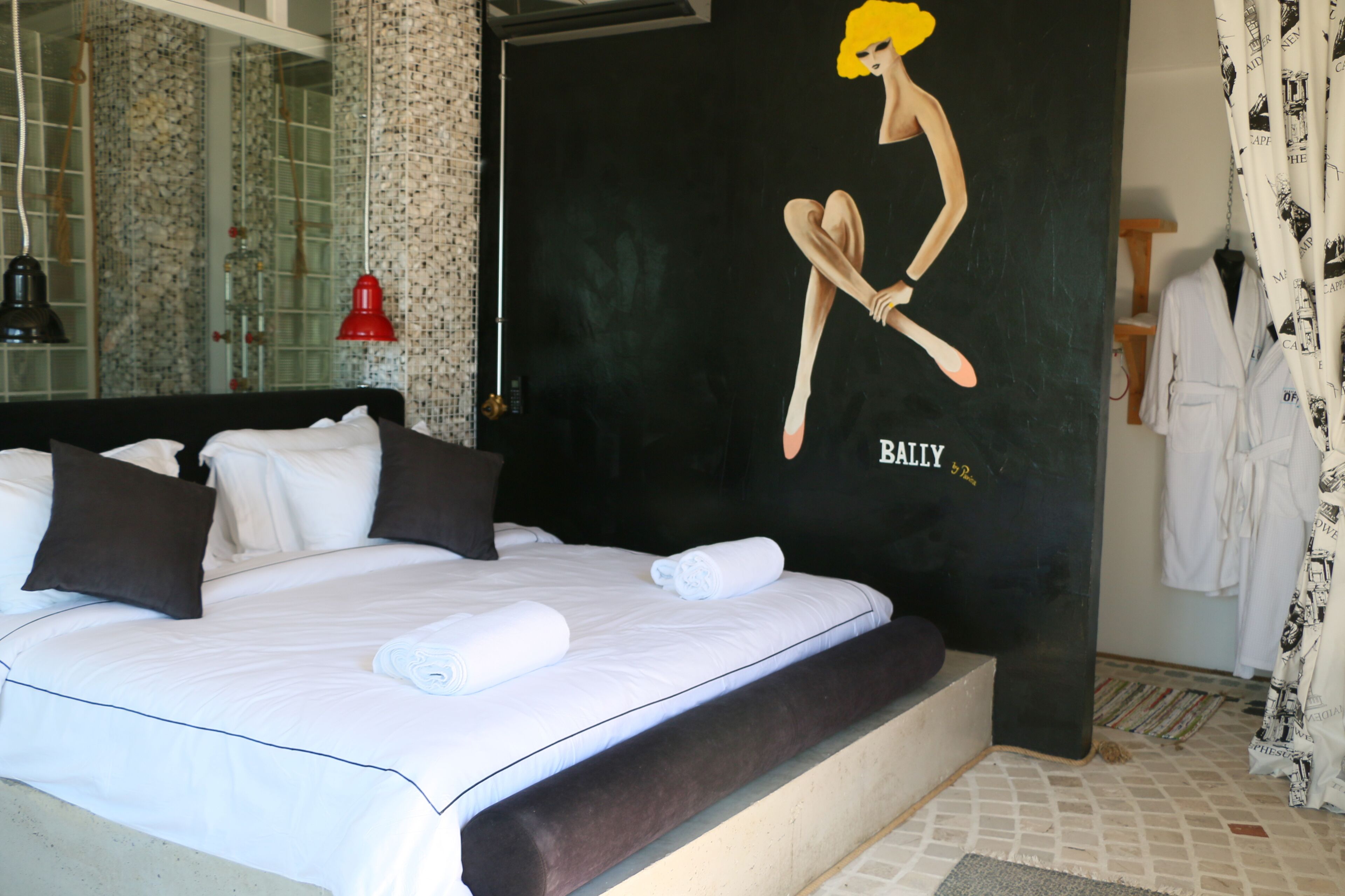 A neatly made bed with white linens and dark pillows, a black wall with a mural, a red pendant light, and a white robe hanging on a rack.