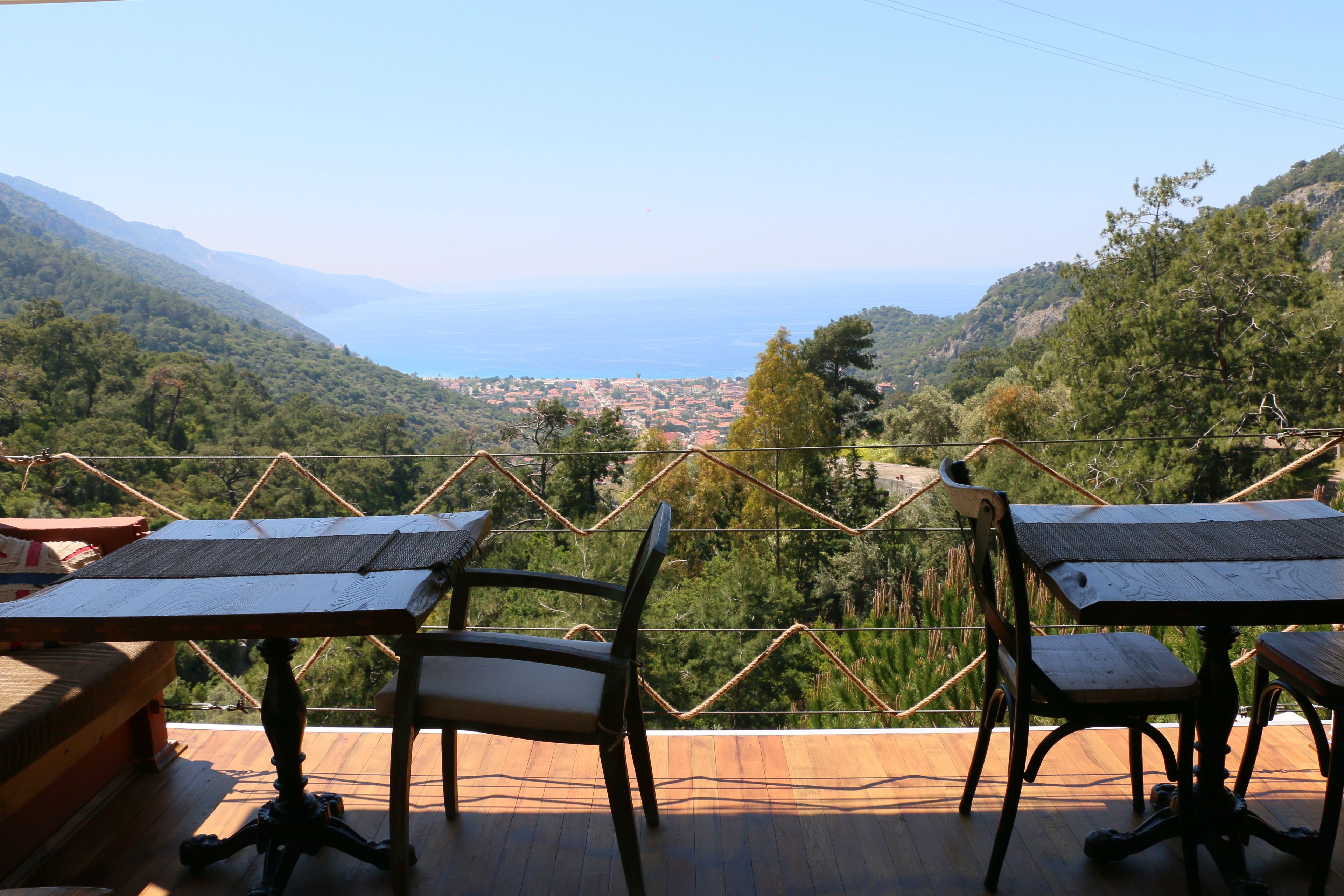 A terrace with tables and chairs overlooking a mountainous landscape and a distant view of the sea.