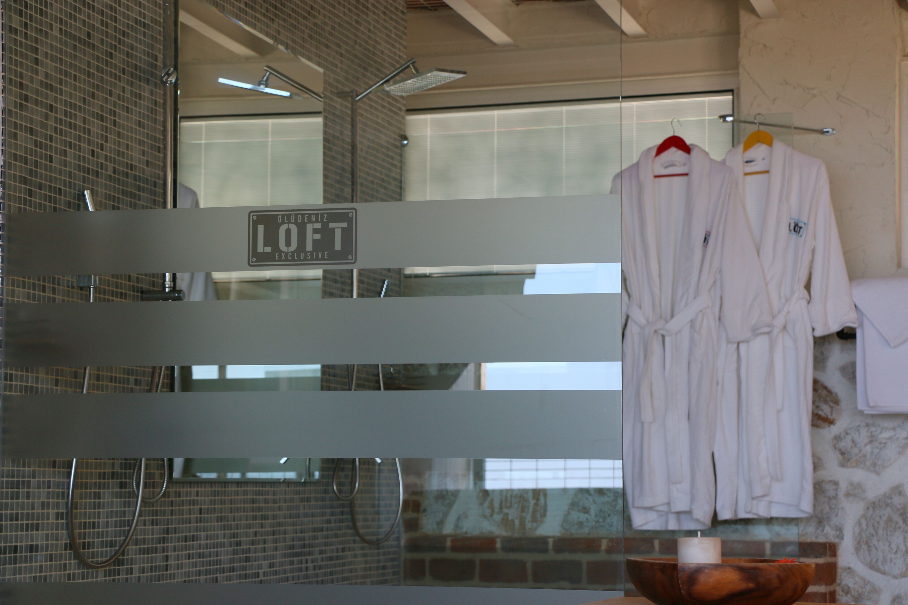 A bathroom with a glass partition, a sign reading "LOFT EXCLUSIVE", and two white robes hanging on a rack.