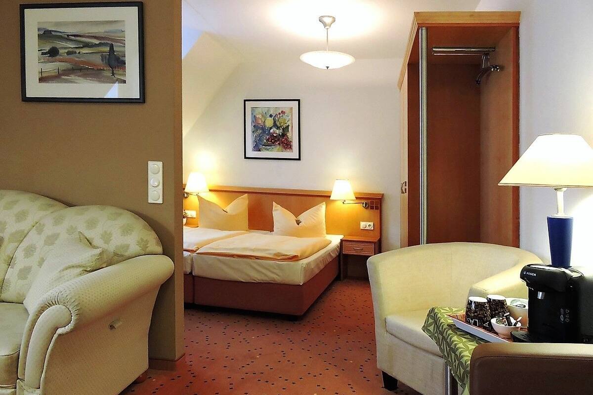 Ein Hotelzimmer mit einem Bett, zwei Sesseln, einem Schreibtisch mit Lampe, einer Kaffeemaschine und Kunstwerken an den Wänden.