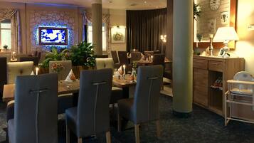 A hotel dining area with a television, dining tables, chairs, and a kitchen area.
